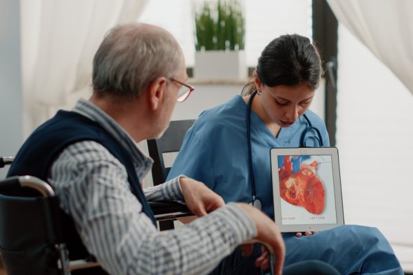 Nurse holding tablet with heart figure for cardiology diagnosis, showing cardiovascular issues to disabled patient at facility. Retired man sitting in wheelchair looking at blood vessel