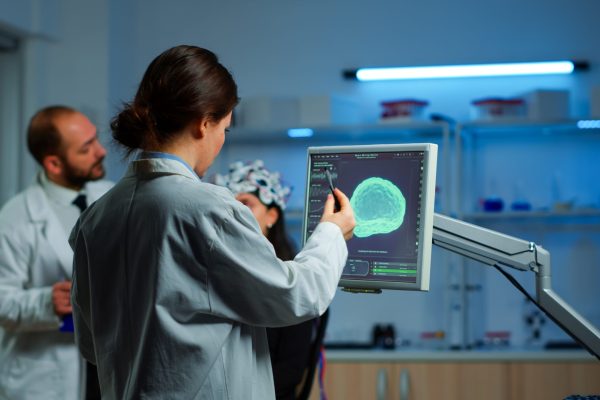 Researcher looking at monitor analysing brain scan while coworker discussing with patient in background about side effects, mind functions, nervous system, tomography scan working in laboratory